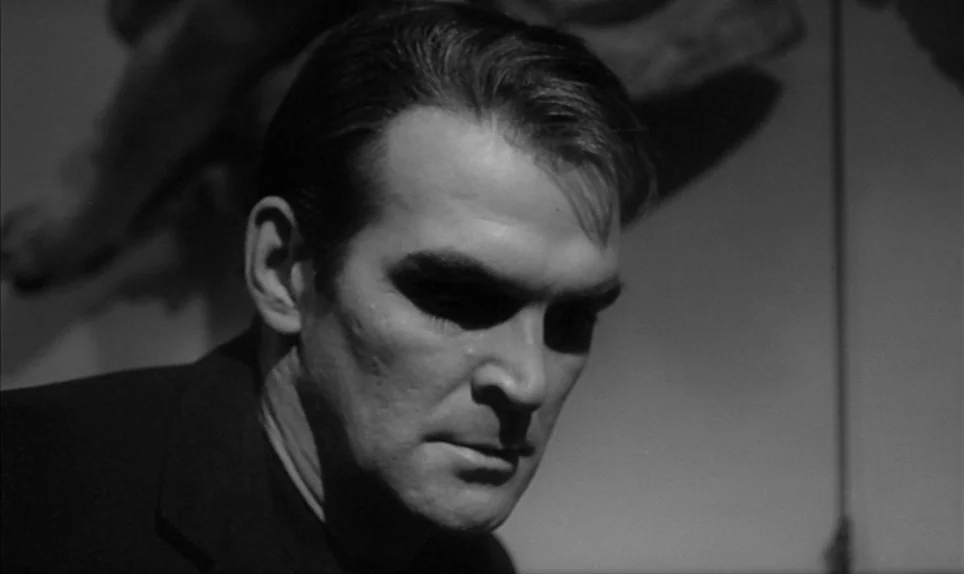 Eva (1962) movie still: closeup — The man sits in a dimly lit indoor environment with deep shadows. He gazes downward with a stoic expression while…