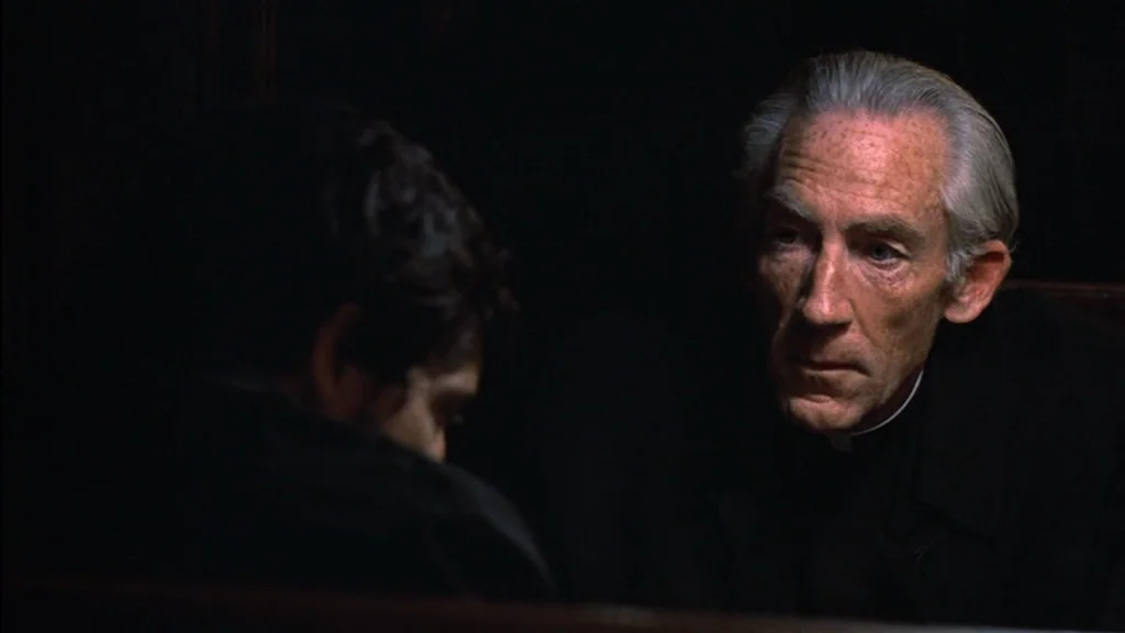 The Exorcist (1973) movie still: medium — A dark, cramped wooden stall with minimal lighting and heavy shadows. An older priest gazes intently…