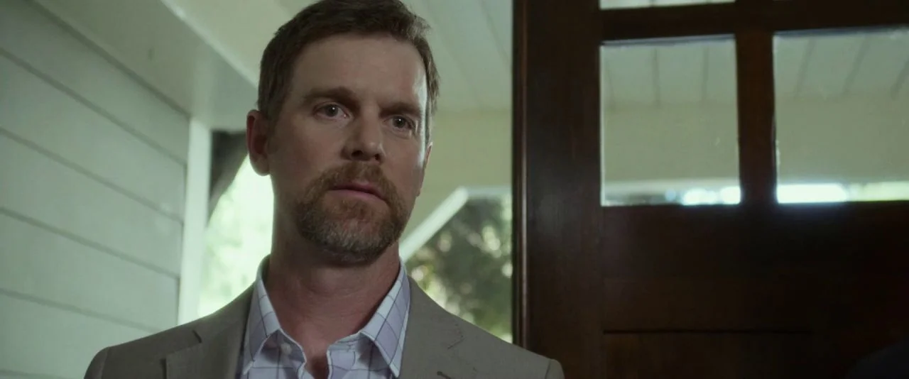 Night Owls (2015) movie still: closeup — A man stands indoors near a wooden door frame and a white paneled wall. He is looking off-camera to his…