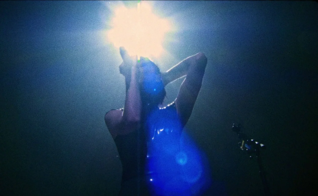 The Nowhere Inn (2020) movie still: medium — A person stands on a dark stage with a bright overhead light source. The subject has both hands behind…