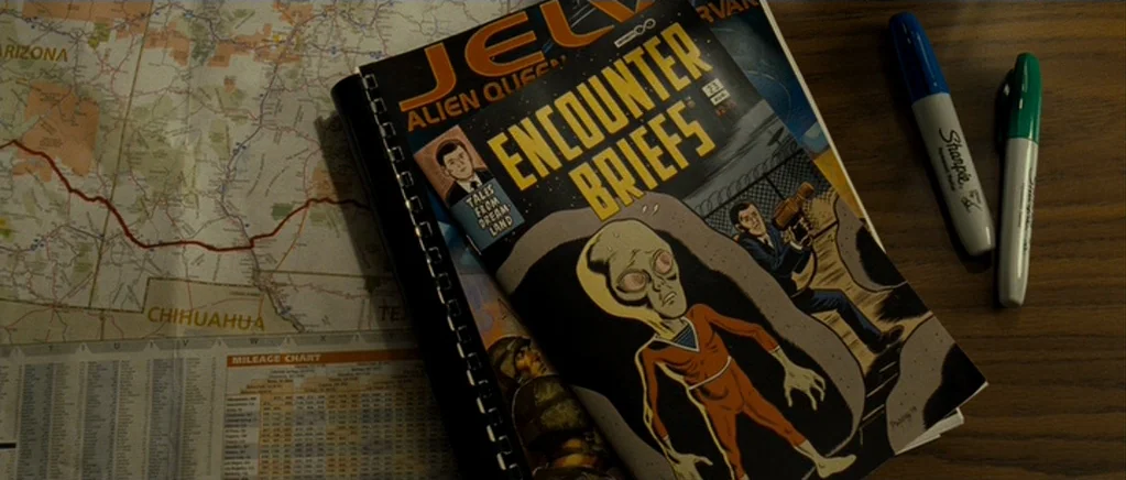 Paul (2011) movie still: closeup — A wooden desktop holds an unfolded road map and a comic book featuring an alien. Two markers, one blue and one…