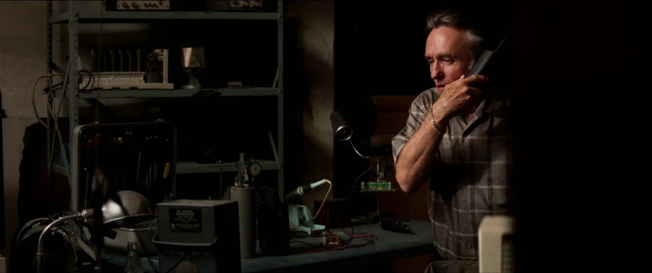 Speed (1994) movie still: medium — A dark workshop cluttered with electronic equipment, metal shelving, and various mechanical tools. A middle-aged…
