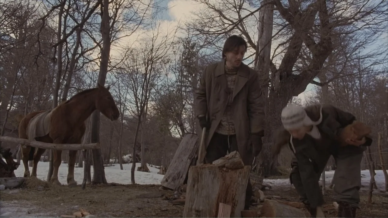 A Distant Place (2020) movie still: fullbody — A snowy, sparse woodland clearing features bare trees and patches of melting ground snow. A man…
