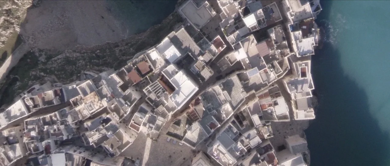 Spring (2014) movie still: establishing — A dense cluster of Mediterranean-style buildings sits on a cliff overlooking the ocean. The white rooftops…