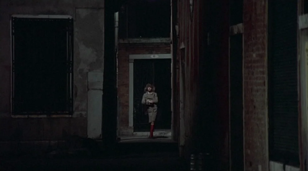Don’t Look Now (1973) movie still: wide — A narrow dark alleyway with aged brick walls and a single doorway in back. A woman in a tan coat and red…