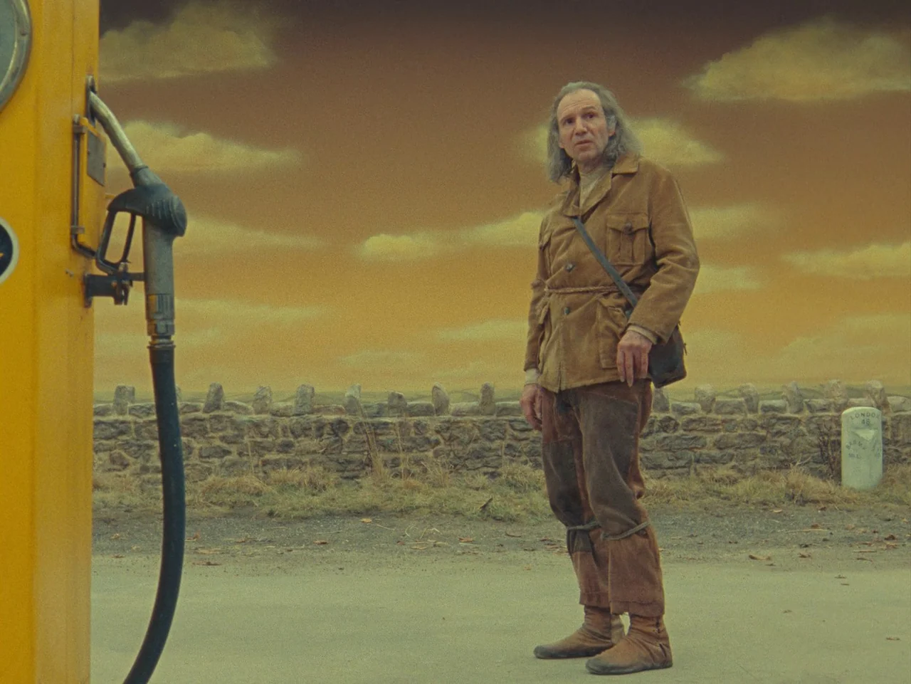 The Rat Catcher (2023) movie still: fullbody — A man stands on asphalt in front of a painted yellow backdrop sky. A vintage gas pump is in the…