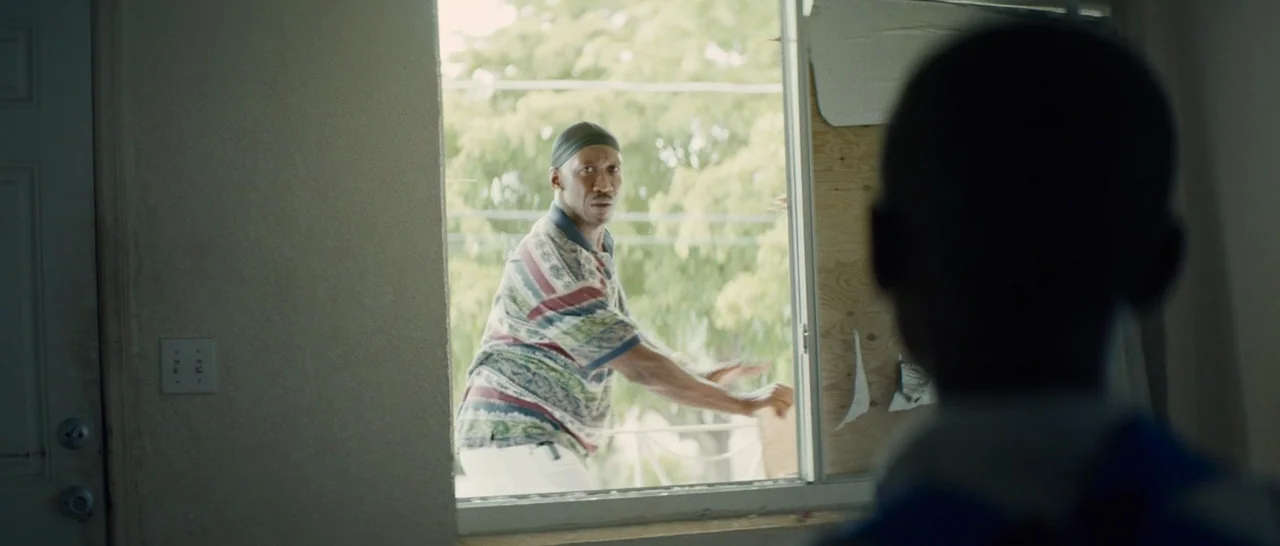 Moonlight (2016) movie still: medium — A man outside stands at a partially opened window of a sparse room. The man reaches into the opening while…
