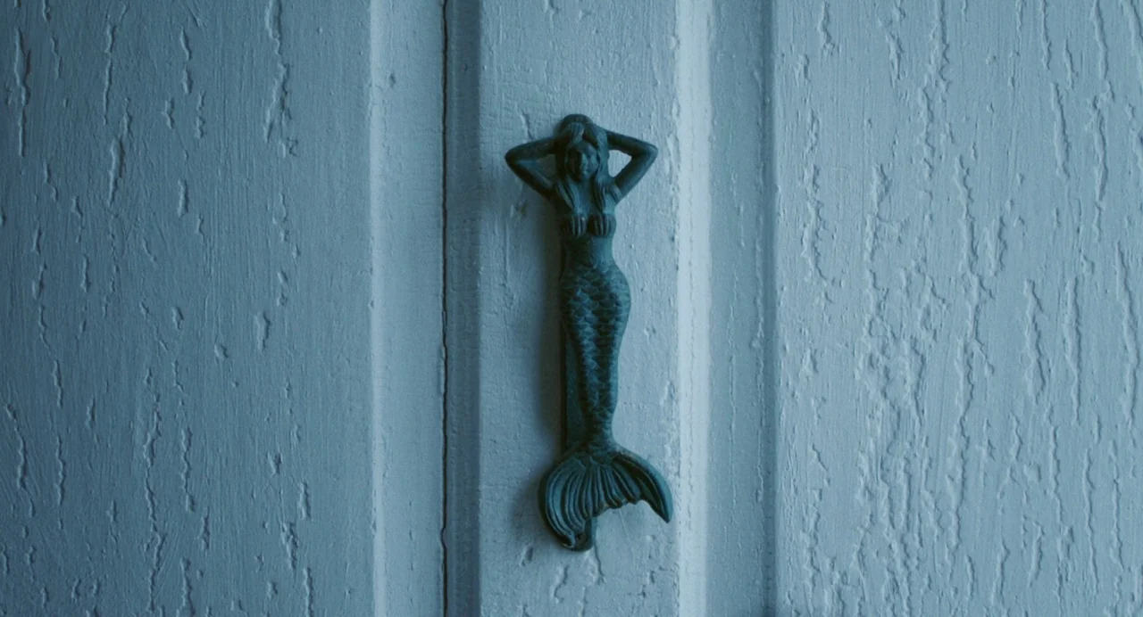 Blow The Man Down (2019) movie still: closeup — A dark metal mermaid door knocker is mounted on a white, textured door. The door surface has…