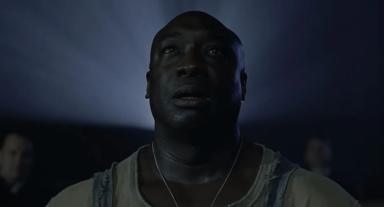 The Green Mile (1999) movie still: closeup — A dark indoor space with a dim light source cast upon a man. A muscular bald man looks upwards while…