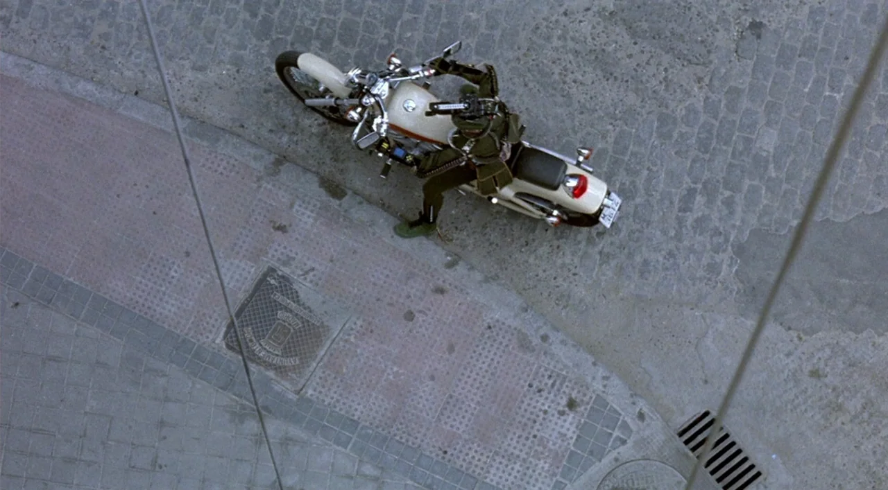 Kika (1993) movie still: wide — A top-down view shows a motorcycle parked on a grey cobblestone street corner. The rider sits stationary on the bike…