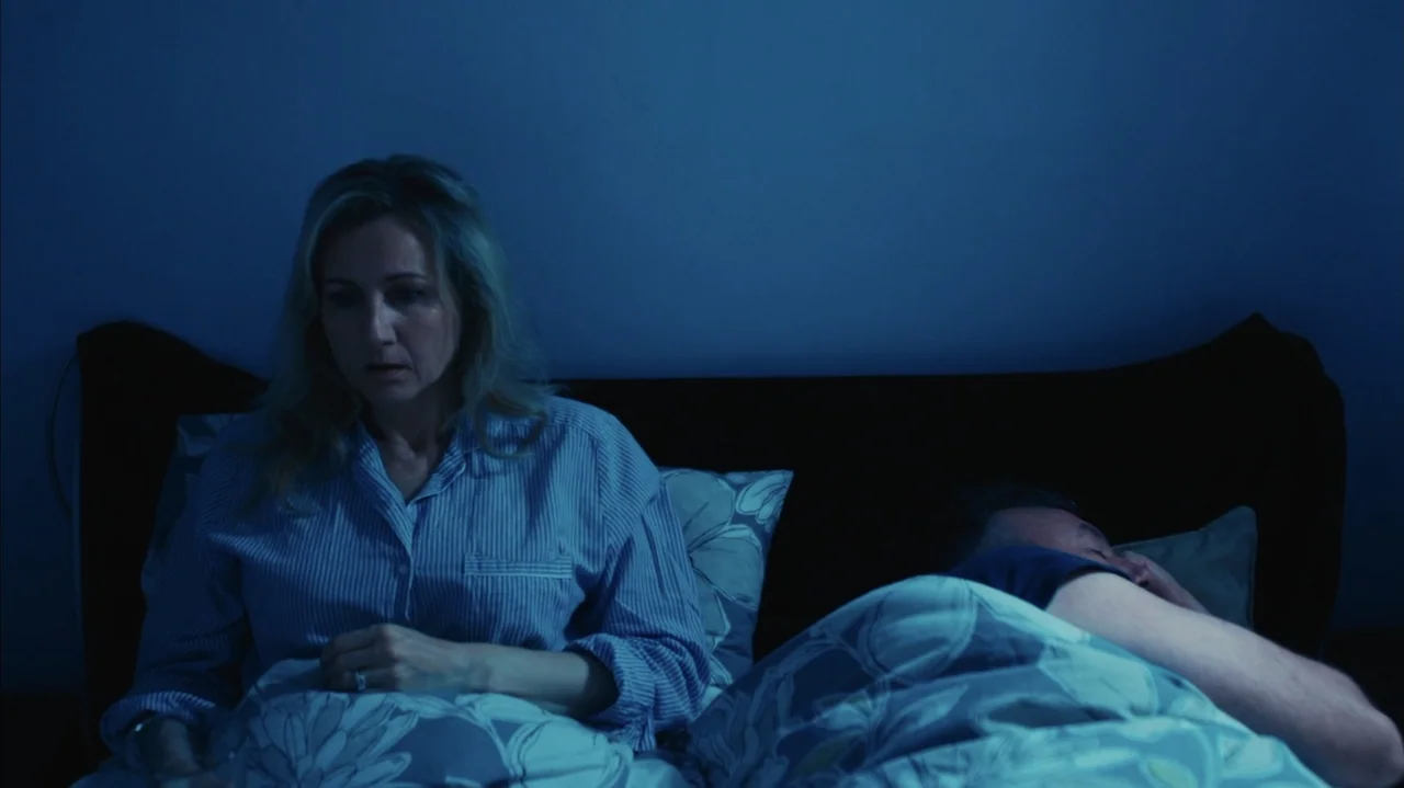 You Might Get Lost (2021) movie still: medium — A dark bedroom with a couple in a bed at night. The woman sits awake while the man remains asleep…