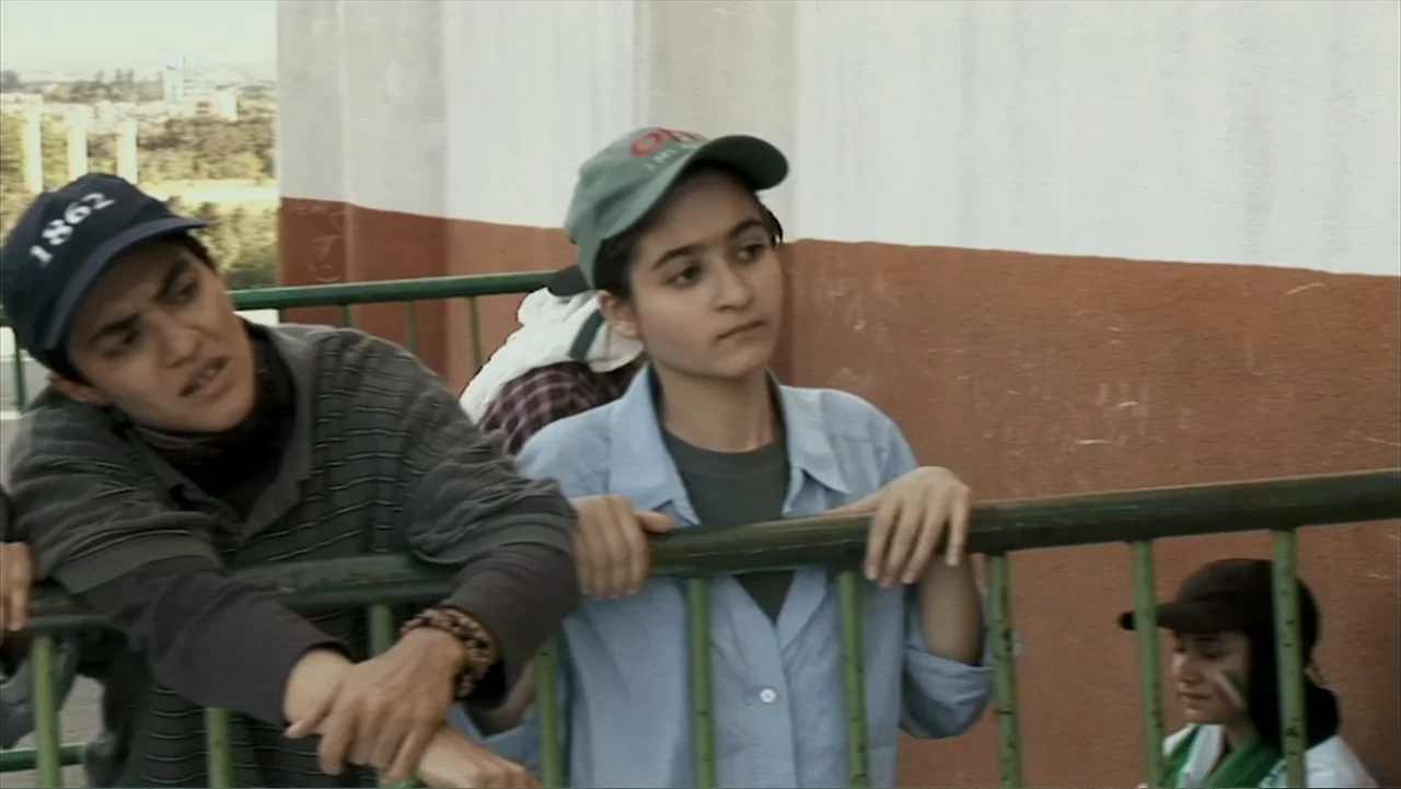 Offside (2015) movie still: medium — An outdoor area with a green metal railing against a pale orange and white wall. Two young people stand near…