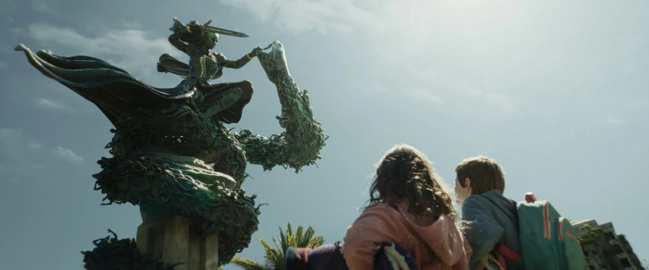 Boy Kills World (2023) movie still: wide — A sunny public plaza features a massive, detailed bronze statue of a sword-wielding figure. Two young…