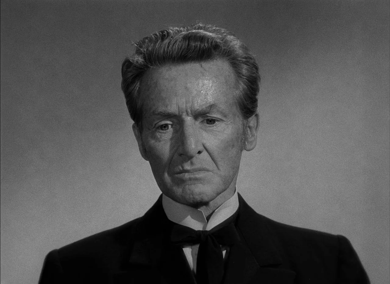 High Noon (1952) movie still: closeup — A plain gray studio background frames an older man from the chest up. The man wears a formal suit and looks…