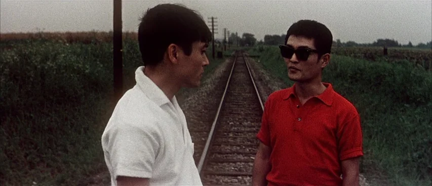 Jigoku (1960) movie still: medium — Two young men stand on gravel railway tracks flanked by grassy fields under overcast skies. The men face each…
