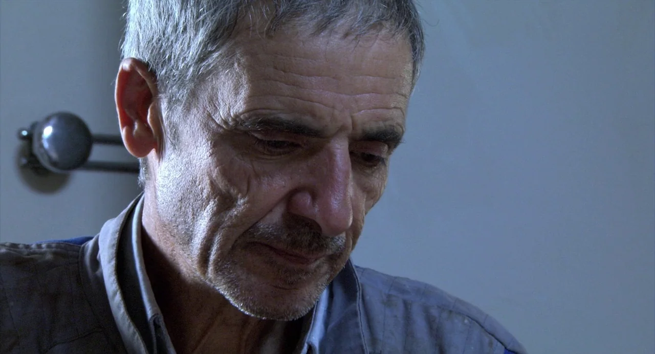 The Secret of the Grain (2007) movie still: closeup — A plain interior room with a metal door handle visible on a grey wall. An elderly man with…