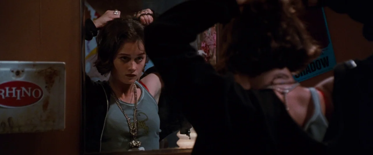 Empire Records (1995) movie still: medium — A dimly lit space features a mirror reflection of a young woman with short hair. The woman looks into…