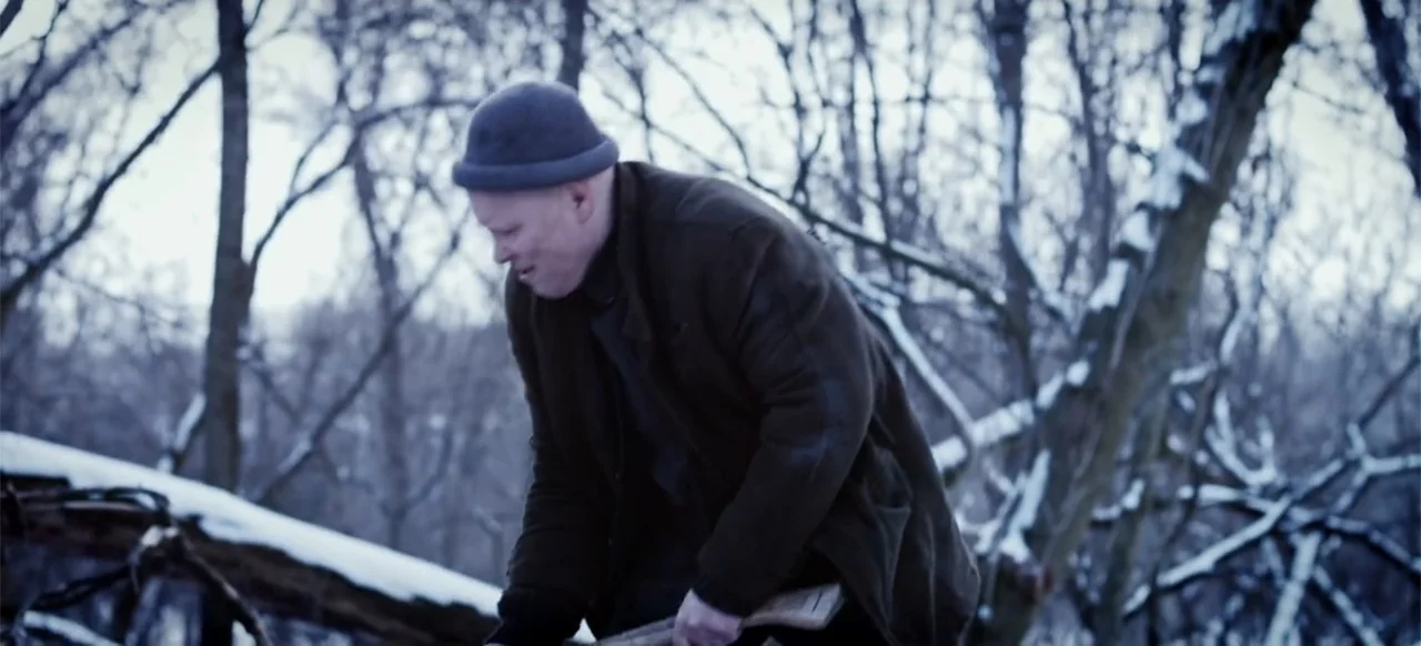 The Russian Bride (2019) movie still: medium — A snowy wooded area contains bare trees and fallen logs under an overcast sky. A man in a dark coat…