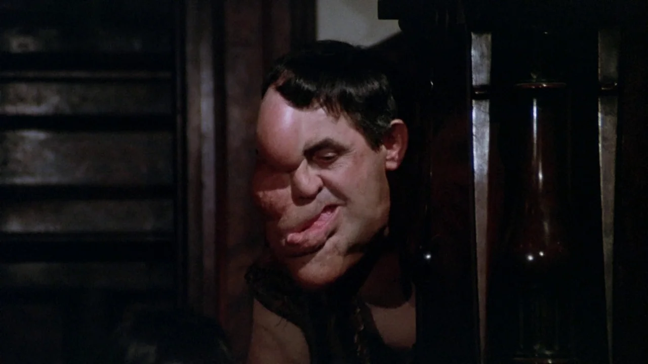 The Sentinel (1977) movie still: closeup — A man with heavy prosthetic facial deformities stands in a dark indoor space. The figure looks toward the…