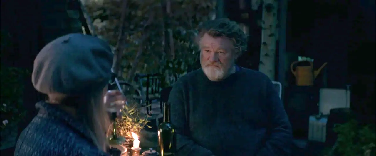 Hampstead (2017) movie still: medium — A dark outdoor patio at night features a man and woman at a table. They are seated across from each other…