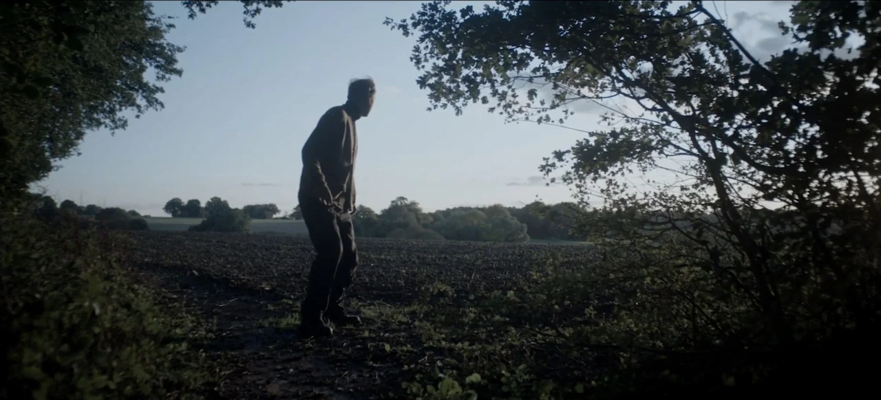 We Go In at Dawn (2020) movie still: wide — An expanse of dark plowed earth stretches toward a distant line of trees. A lone man in dark clothing…
