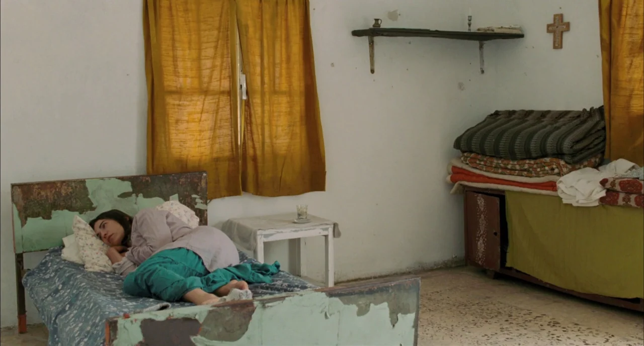 Incendies movie still: wide — A spartan room features peeling painted furniture and simple mustard curtains over a window. A woman lies curled up…