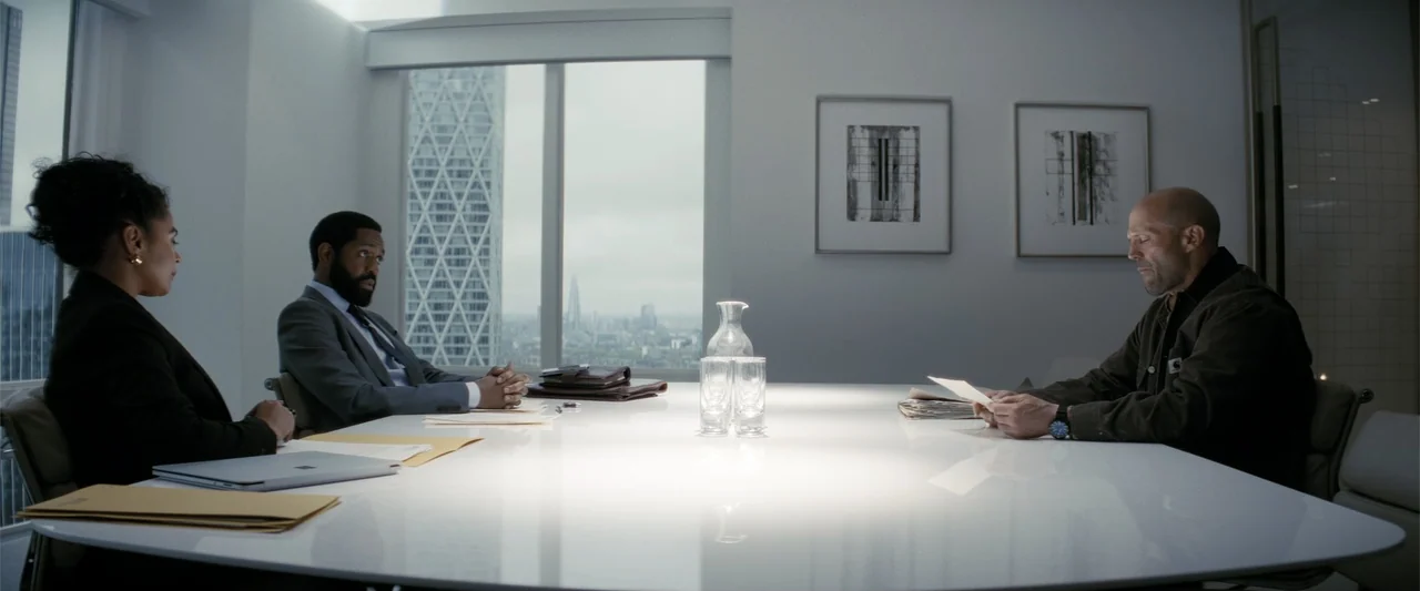 A Working Man (2025) movie still: medium — A modern white conference room features a large window with a distant city skyline. Three people sit…