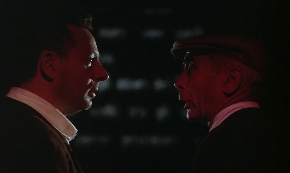 Cinema Paradiso (1988) movie still: medium — A dark, minimally lit interior space provides the backdrop for two men facing profile. The two men…