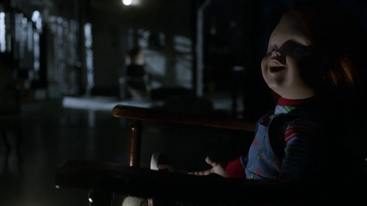 Curse of Chucky (2013) movie still: medium — A dark nursery room features a wooden rocking chair in the foreground. A Chucky doll sits in the chair…