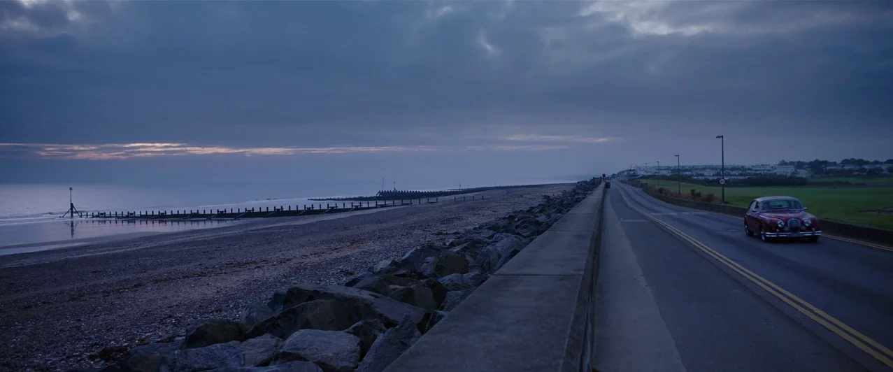 Ghost Stories (2017) movie still: establishing — An asphalt road runs parallel to a shingle beach under a dark overcast sky. A red classic car…