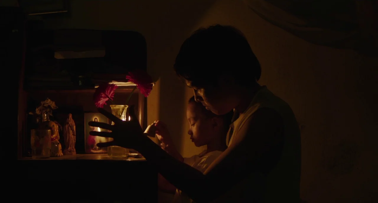 Inside The Yellow Cocoon Shell (2023) movie still: medium — A dimly lit indoor space features a small religious altar with flowers and figurines. An…