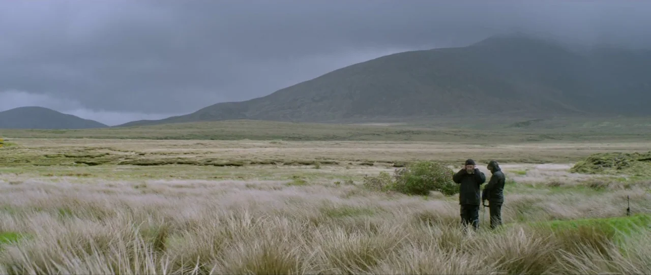 Silence (2012) movie still: wide — A vast, grassy bog landscape stretches toward a large, cloud-covered mountain in the distance. Two men in outdoor…