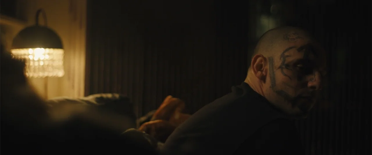 Skin (2009) movie still: medium — A dimly lit room with a man sitting on a couch in the foreground. The man has prominent tattoos on his face and…