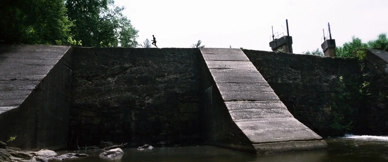 The Hunger Games (2012) movie still: wide — A long, weathered concrete dam spans across a stream under a bright sky. A lone figure in athletic…