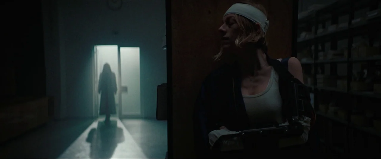 Cuckoo (2024) movie still: medium — A dimly lit storage room with metal shelving units lines the wall in shadow. A woman in a medical robe hides…