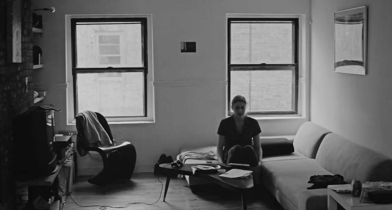 Frances Ha (2012) movie still: wide — A sparse modern apartment features a brick wall, two windows, and a sofa. A woman sits on the sofa with her…