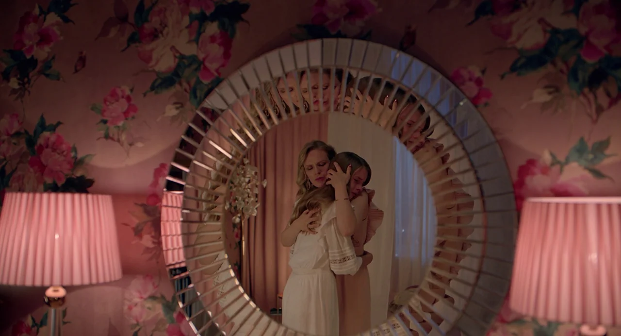 Hatching (2022) movie still: medium — A bedroom with floral wallpaper and two pink bedside lamps frames a circular mirror. Two women in light…