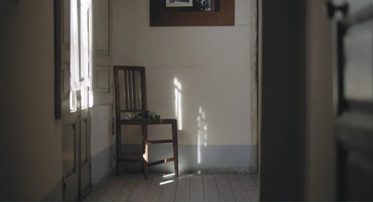 Los días azules (2025) movie still: medium — A light-colored hallway wall features a wooden chair with a plant resting on it. Natural light casts…