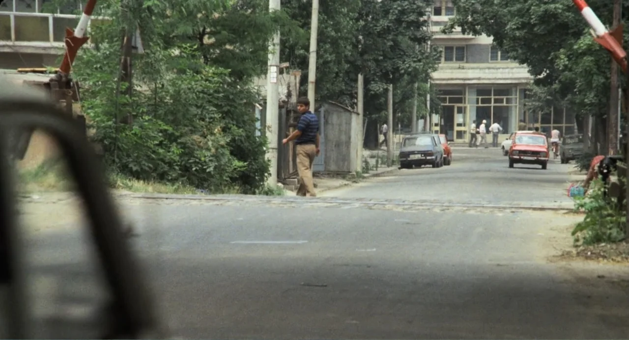 Do Not Expect Too Much from the End of the World (2023) movie still: wide — A suburban road crosses an active railway track marked by a lowered…