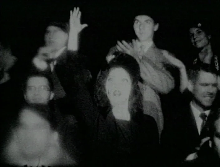 Cowards Bend The Knee (2003) movie still: medium — A group of people sit in a dark theater looking toward the camera. Multiple individuals raise…