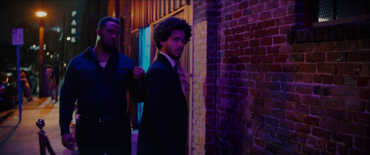 Night Teeth (2021) movie still: medium — A city street at night features a weathered red brick wall bathed in neon. Two men in formal attire stand…