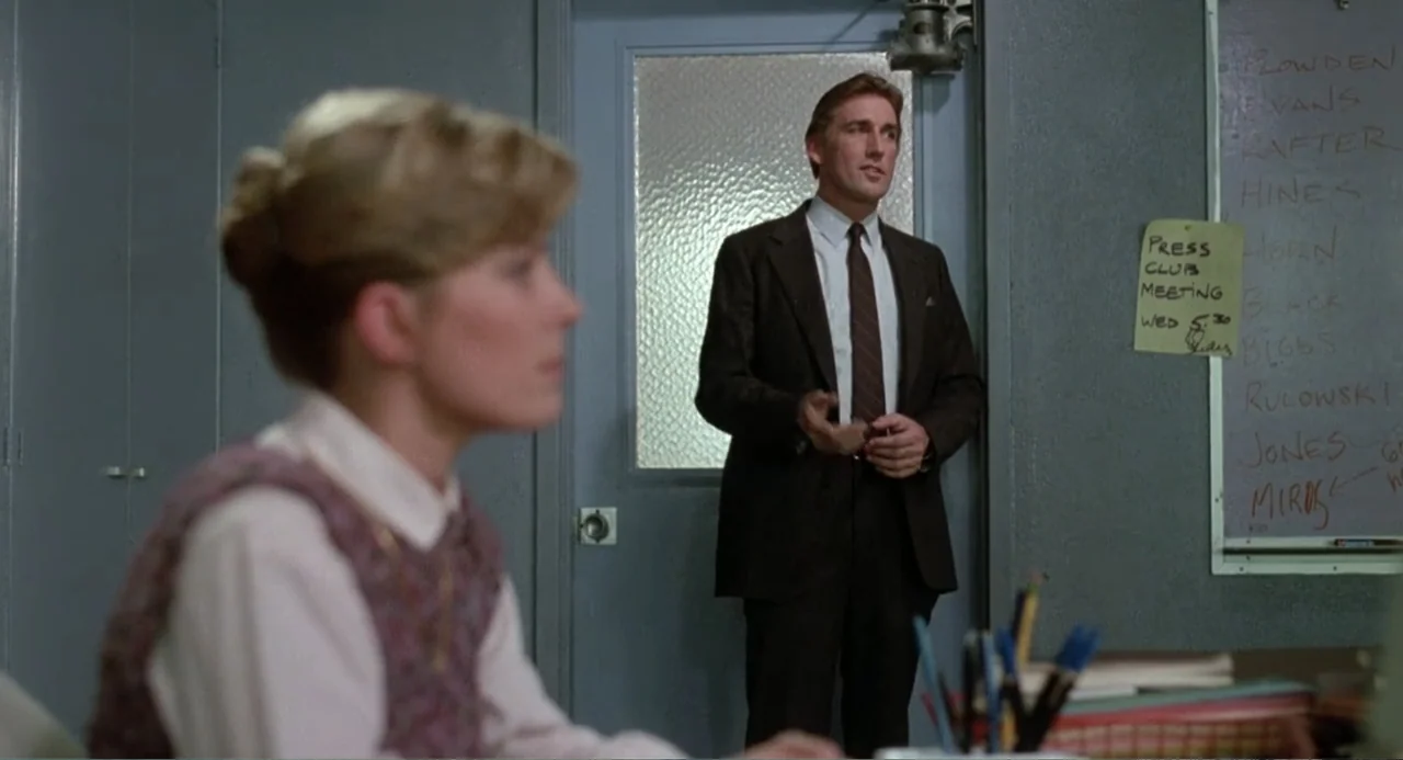 Dangerously Close (1986) movie still: medium — A utilitarian office features gray walls and a notice board with handwritten text. A man in a…