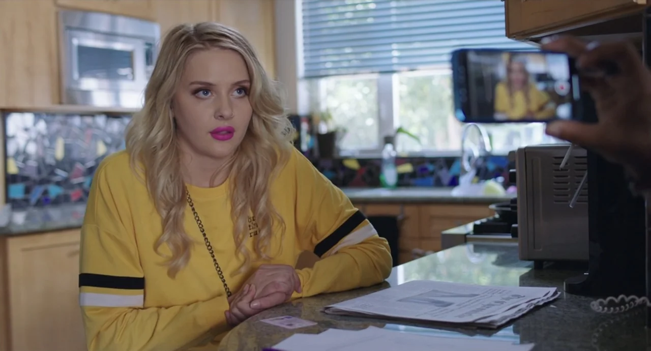 Pleasure (2021) movie still: medium — A kitchen features light wood cabinets and a granite countertop with a newspaper. A young woman with blonde…