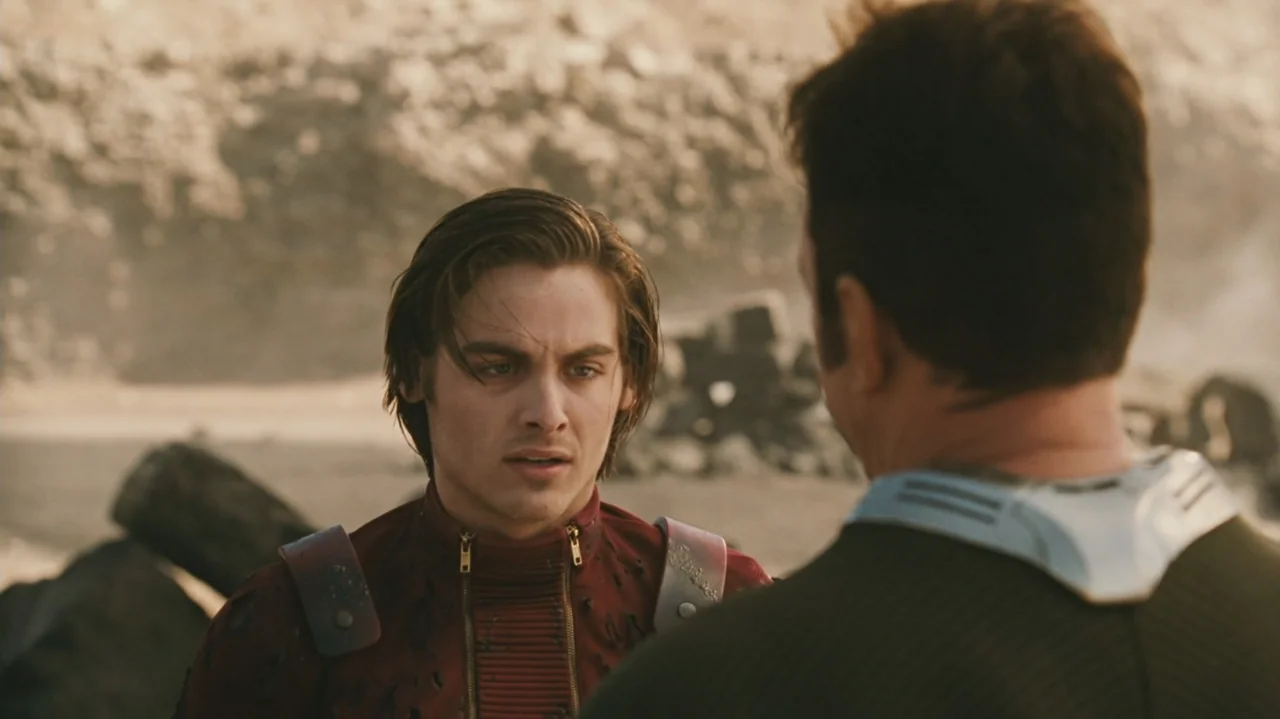 Zoom (2006) movie still: medium — A dry, rocky terrain serves as the backdrop for two men in futuristic gear. A man in a maroon suit speaks to an…