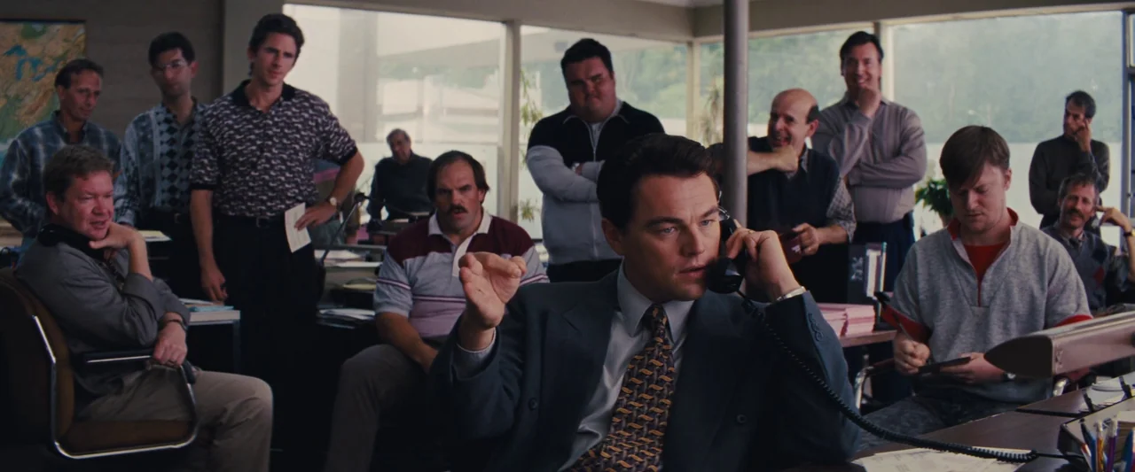 The Wolf of Wall Street (2013) movie still: medium — An open plan workspace features large windows and groups of men at desks. A man in a suit sits…