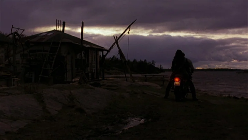 Dance Me Outside (1994) movie still: wide — A dark rocky shoreline features a small cabin with a leaning ladder and primitive crane. A lone person…