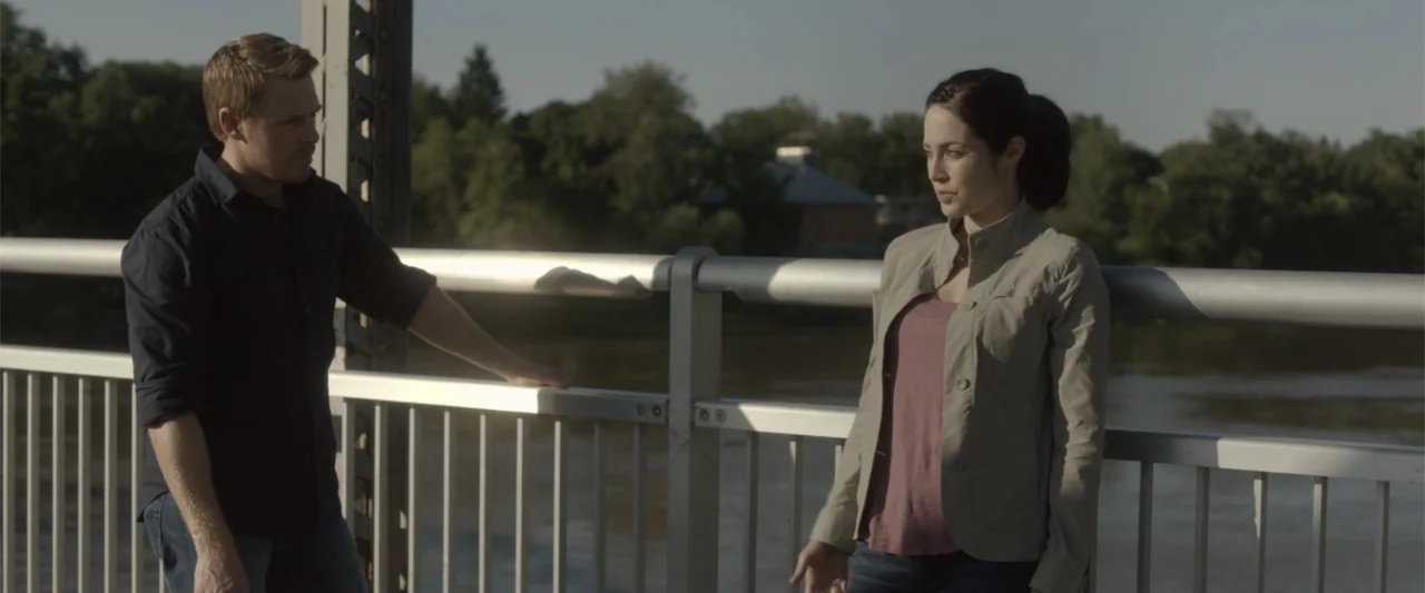 Radius (2017) movie still: medium — A man and woman stand on a metallic bridge over a river during sunset. The subjects face each other while…