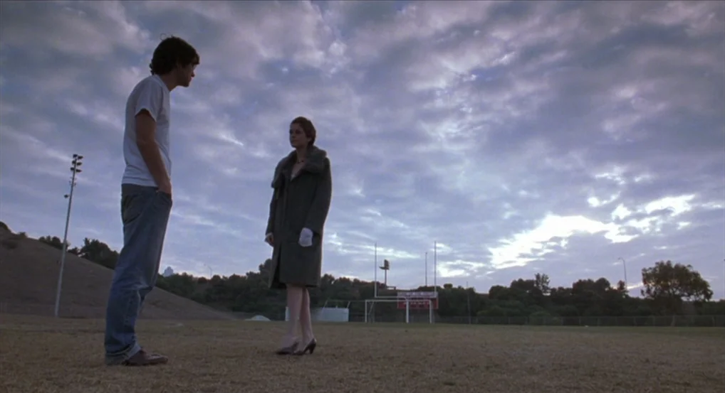Brick (2005) movie still: fullbody — An open field at twilight features a goalpost against a cloudy, dim sky. A man in a white shirt and a woman in…