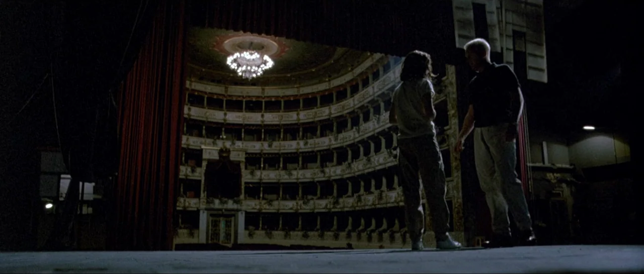 Opera (1987) movie still: fullbody — An expansive historic Italian-style auditorium with multiple tiers of boxes faces a dark stage. A man and a…
