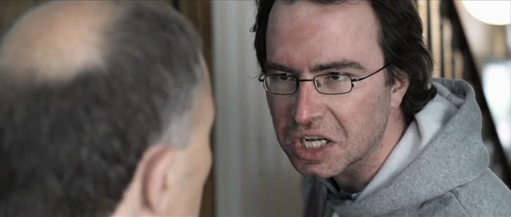 Down Terrace (2009) movie still: medium — A narrow residential hallway with neutral-colored walls provides the background for this interaction. A…